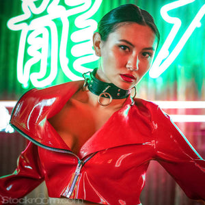 A woman with dark hair sits in front of a glowing green and red neon sign. She is wearing a red latex moto jacket and black latex panties. She is wearing the black leather 3-Ring Slave Collar With A Locking Buckle.