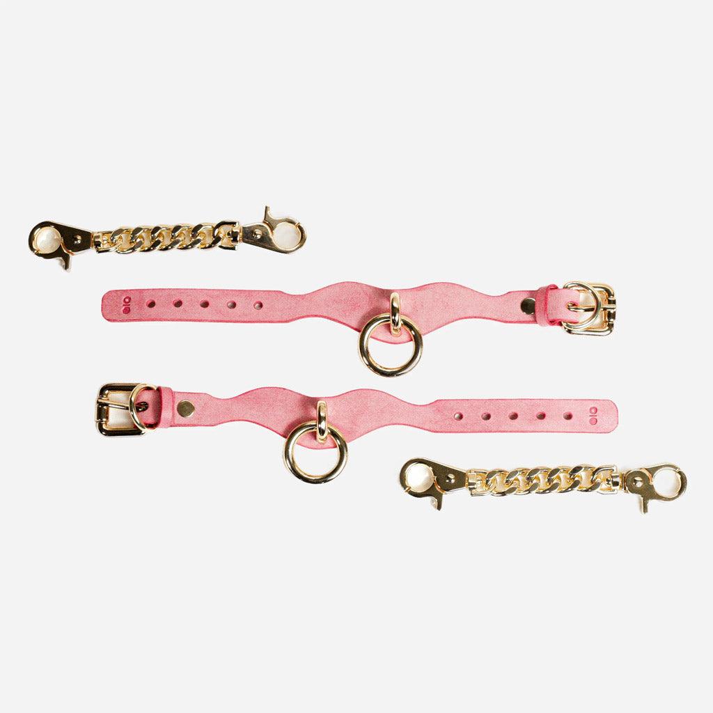 A pair of pink leather BDSM wrist cuffs on a white background, spread out full length. Next to them are two gold-colored chains with lobster-claw fasteners.