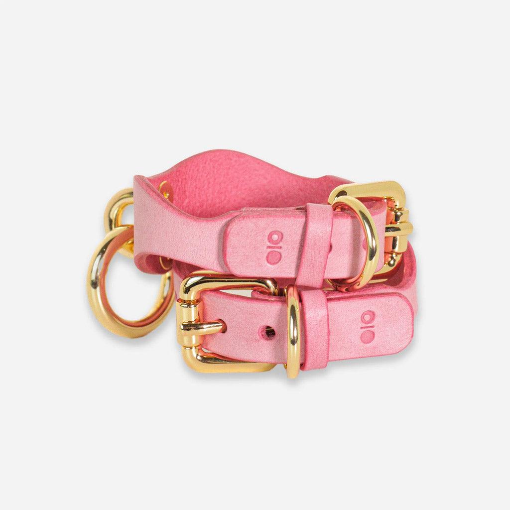A pair of pink leather day cuffs on a white background, one stacked on the other. They're positioned to show the buckling closure mechanism with gold-colored hardware.