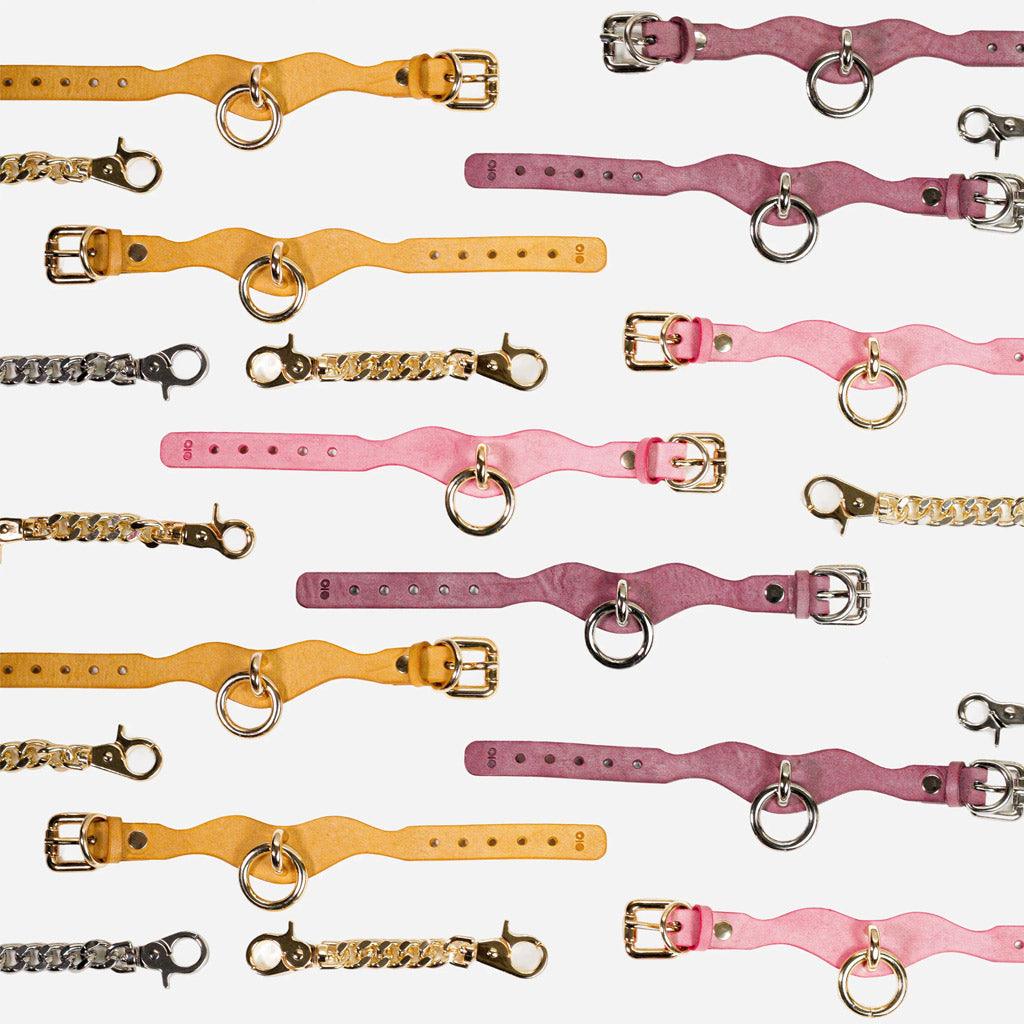 A full square showing several examples of the leather day cuffs open against a white background, with gold and silver connecting chains. The leather is in three colors: bright yellow, lavender, and pink.