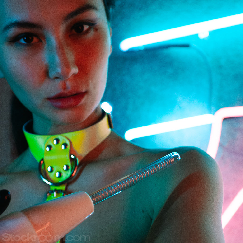 A woman with her dark hair pulled back and wearing a neon yellow bust harness and black latex driving gloves is shown from the bust up. She holds a white neon wand with the KinkLab Coiled Probe Neon Wand Electroplay Attachment.