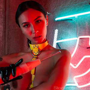 A woman with her dark hair pulled back and wearing a neon yellow bust harness and black latex driving gloves is shown from the bust up. She holds a white neon wand with the KinkLab Coiled Probe Neon Wand Electroplay Attachment.