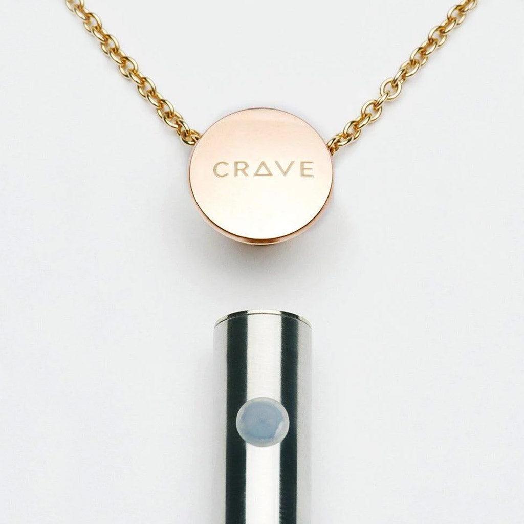 The bottom of the Vesper Pendant Necklace Vibrator is shown below a rose gold chain, which has a pendant with the Crave logo on it. The vibrator has one small button.