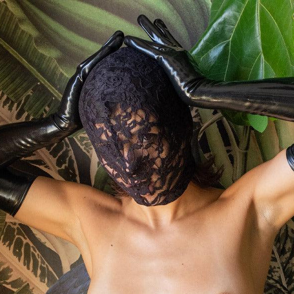 A topless woman wearing black latex opera gloves sits against a background of plants. Her hands are behind her head, and she is wearing the Scandal Lace Hood. The hood completely covers her head and face, except her mouth, and is made of black floral-patterned lace.