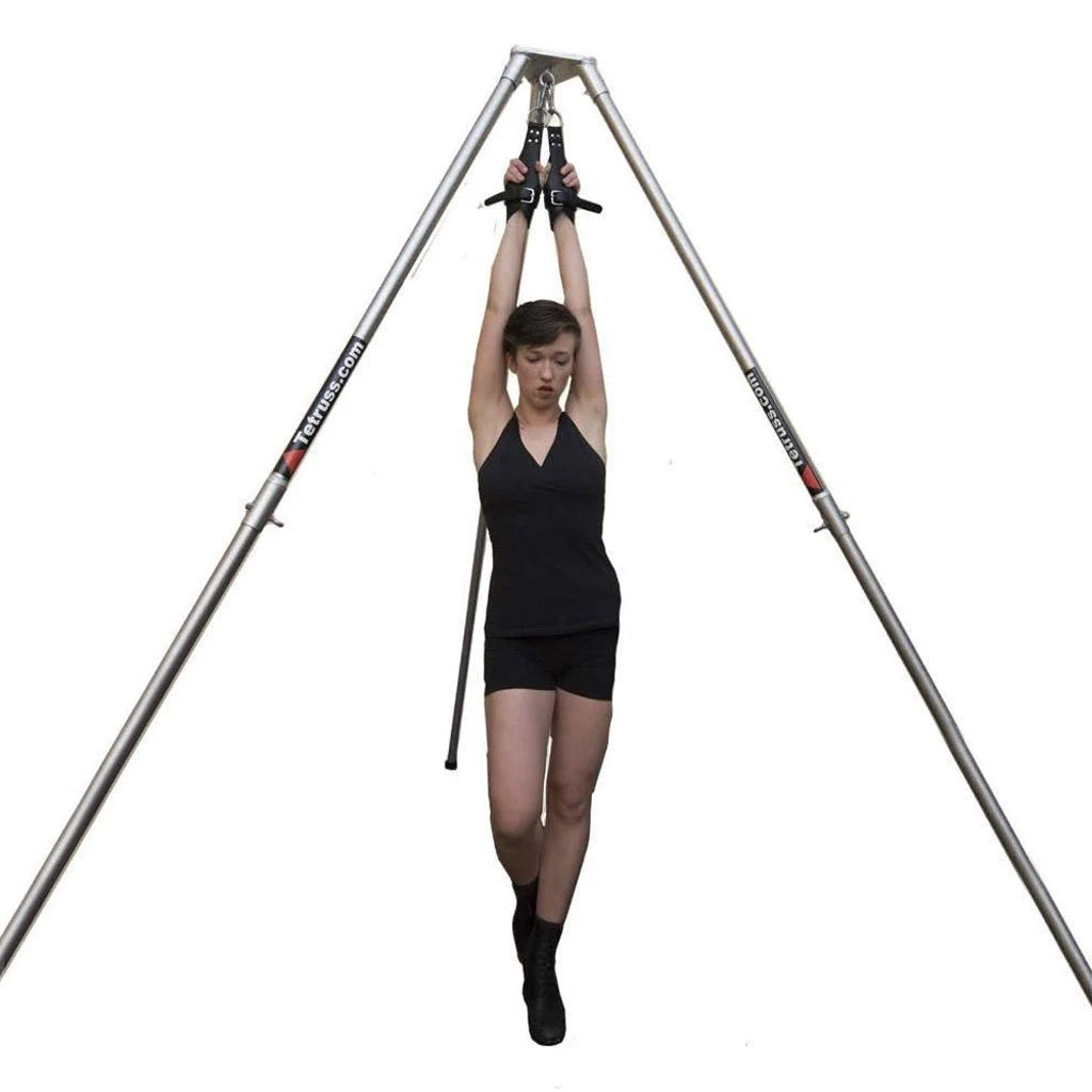 The Tetruss Suspension Bondage Frame, made of 3 silver poles in a triangle formation, is shown in use. A woman in a black tanktop and shorts is wearing black suspension cuffs, which are chained to the top of the frame, lifting her arms above her head.