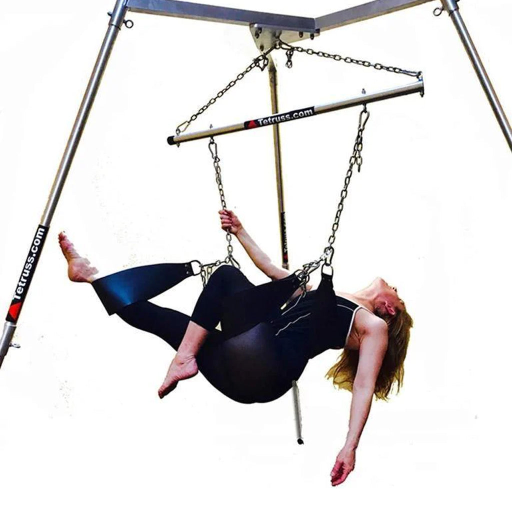 The Tetruss Maxximus Portable Dungeon Deluxe Bundle is shown in use against a blank background. The metal bondage frame has a spreader bar attached to the center, which supports 3 leather stirrups. A woman in a black bodysuit is reclining in the stirrups.