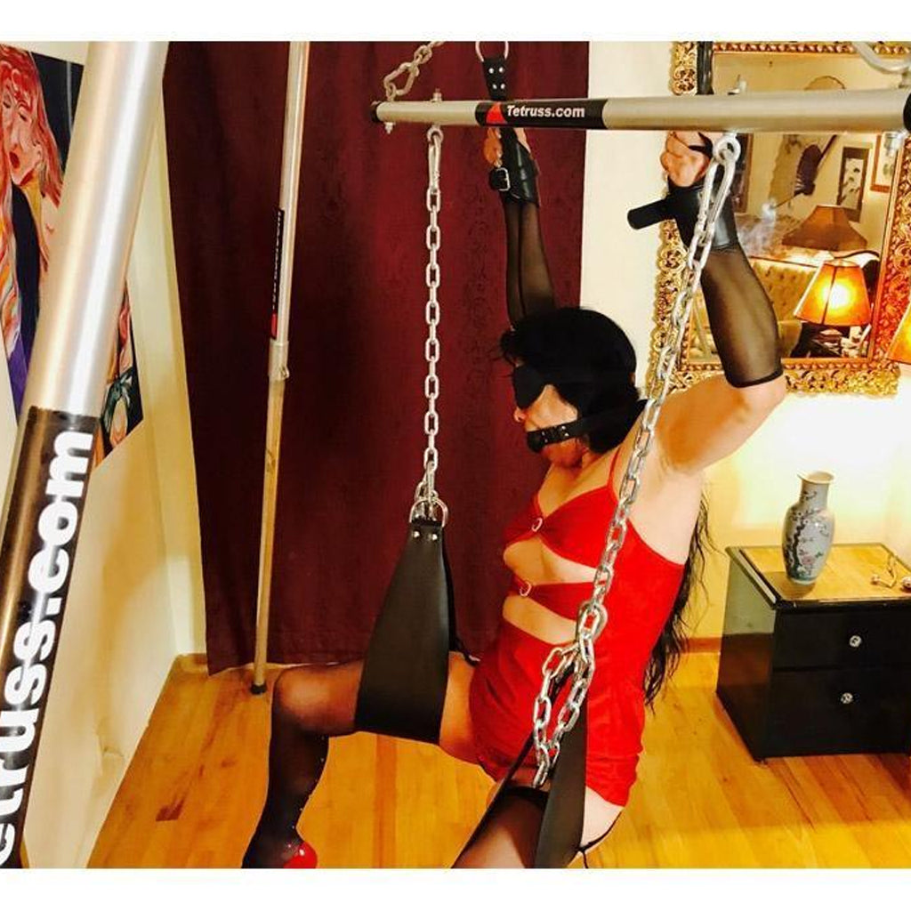 A woman in a red dress is shown using The Tetruss Maxximus Portable Dungeon Deluxe Bundle. She is sitting up with her legs supported by the stirrups, and is holding on to the suspension cuffs she is wearing. She is also wearing a black blindfold and a gag.