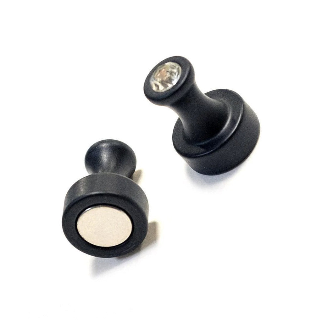 A pair of Magnetic Nipple Power Pinchers are displayed against a blank background. They are black and resemble the back of a thumbtack with a small crystal at the top. There is a magnet at the base of the pinchers.