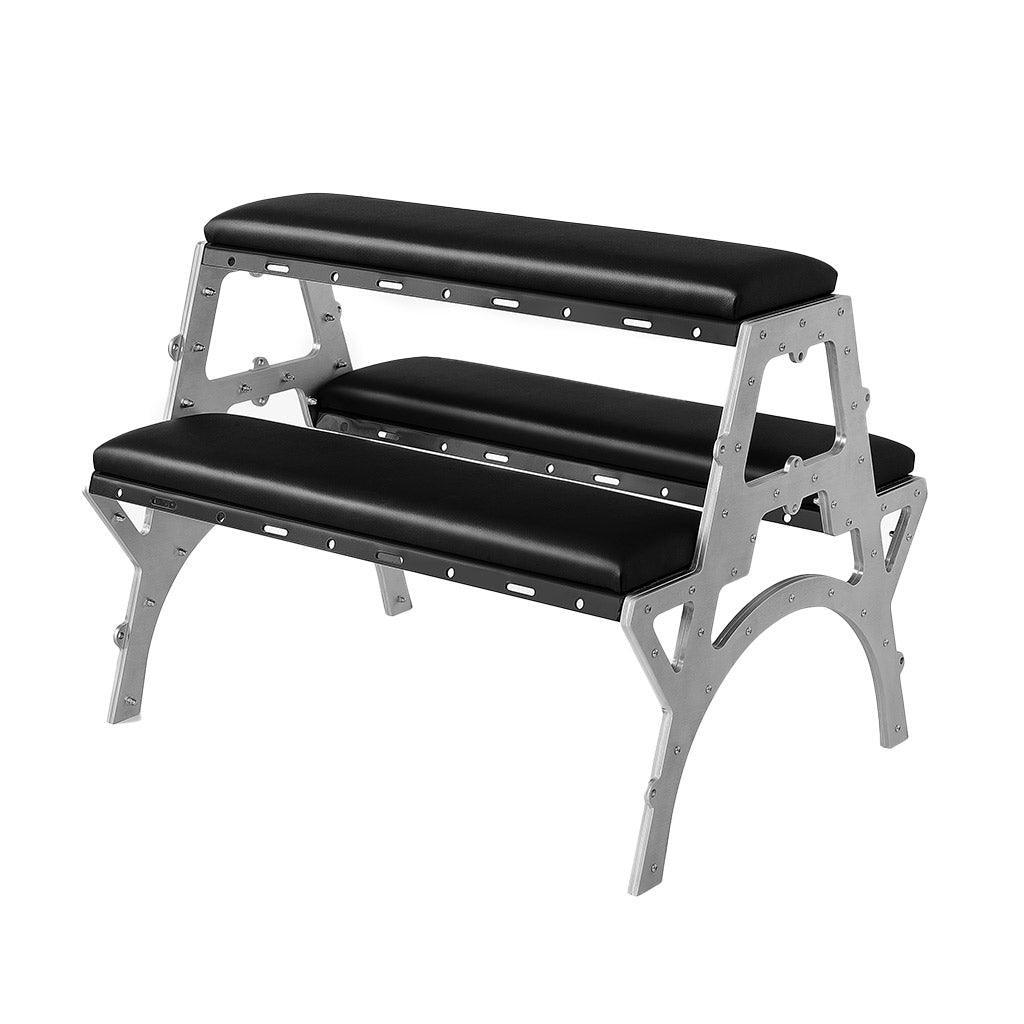 Product shot of the Modern Bondage Spanking Bench on a white background. The frame is made of metal. There are two padded shelves on either side for the person’s knees. A higher shelf between the two of them supports the torso.