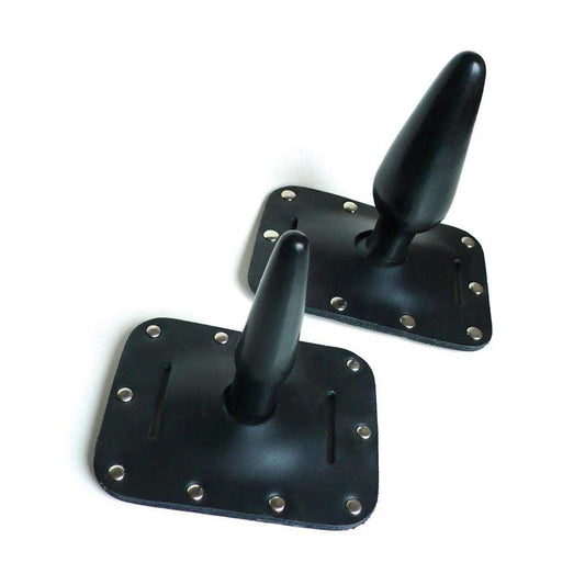 Two Harness Butt Plugs are displayed against a blank background, one small and one large. The plugs are made of black rubber, and the base is embedded in black leather lined with metal rivets.
