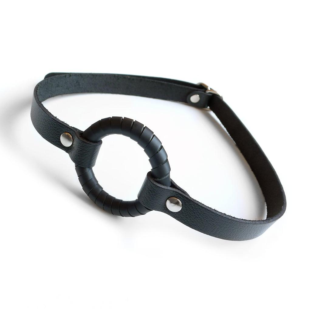 The O-Ring Gag is shown against a blank background. It is a metal O-ring wrapped in a thin strip of black leather attached to a thin, black leather strap.