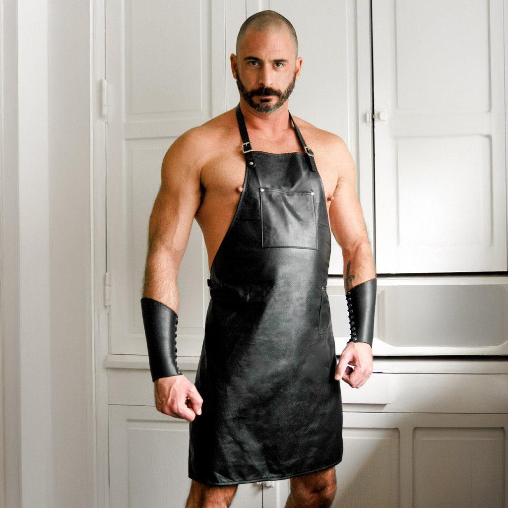 A shirtless muscular man is shown wearing the black Leather Apron, which has adjustable straps and a pocket on the chest. The apron cuts off above the knees. He is also wearing leather arm gauntlets.