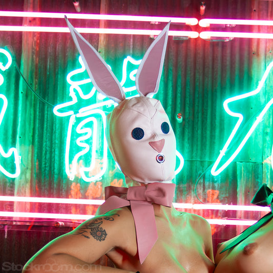 A nude woman reclines in front of a green and red neon sign. She wears the white Leather Bunny Hood, which covers her face and head and ties with a pink bow around her neck. The hood has large ears, blue eyes, a pink nose, and a small opening over the mouth.
