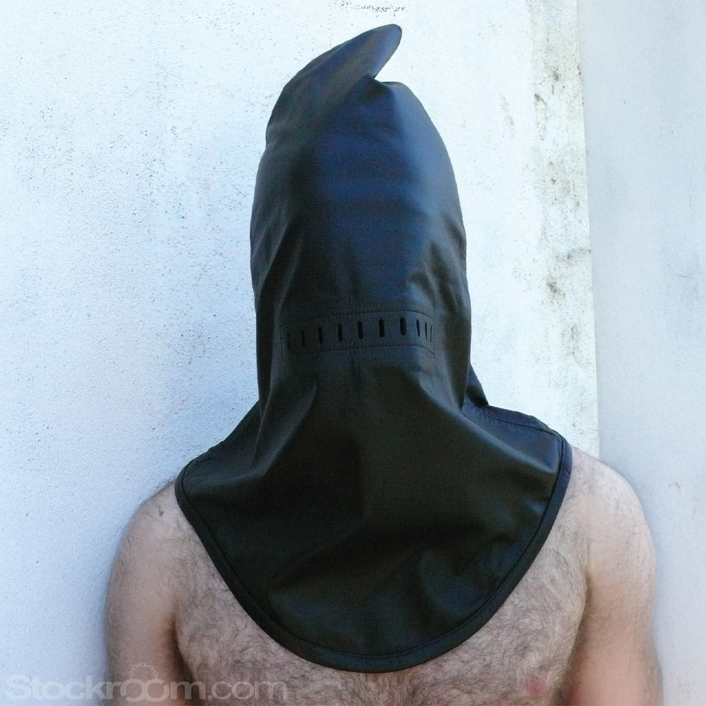 The Leather Guillotine Hood is shown on a man with dark chest hair standing in front of a white wall. The hood fits loosely and has a point at the top and a U-shaped bottom that reaches his chest.