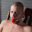 Load image into Gallery viewer, A shirtless man with a buzzed head and facial hair is shown sitting in a wooden chair. He has the Medical Silicone Ball Gag in his mouth.