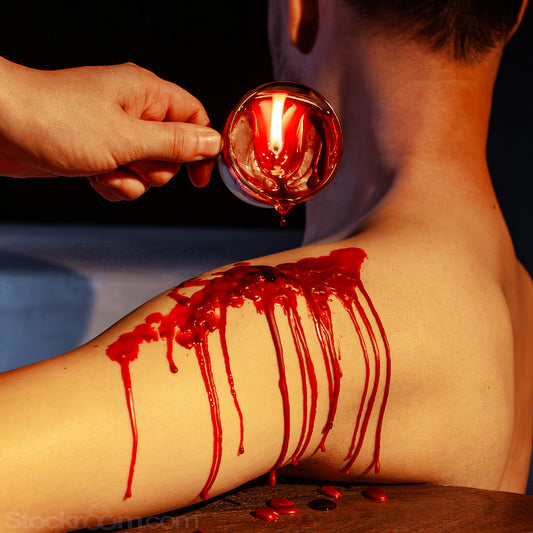 A hand holding the easy pour wax play candle with wooden handle above a person’s shoulder, pouring liquid wax that drips down the skin in glossy streams against a dark background.