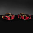 Load image into Gallery viewer, A pair of the Melanie Rose Designs x The Stockroom Wrist Cuffs, made of black and red leather braided together with a gold O-ring in the center, are displayed against a black background.
