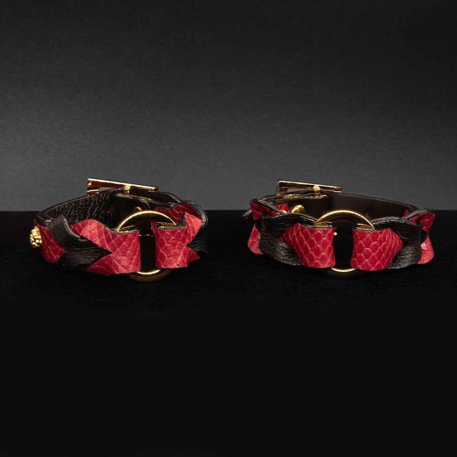 A pair of the Melanie Rose Designs x The Stockroom Wrist Cuffs, made of black and red leather braided together with a gold O-ring in the center, are displayed against a black background.