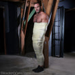 Load image into Gallery viewer, A brunette man with chest hair stands against a wooden beam. He is wrapped from his feet to under his pecs in Clear Latex Bondage Strips, making him resemble a mummy.