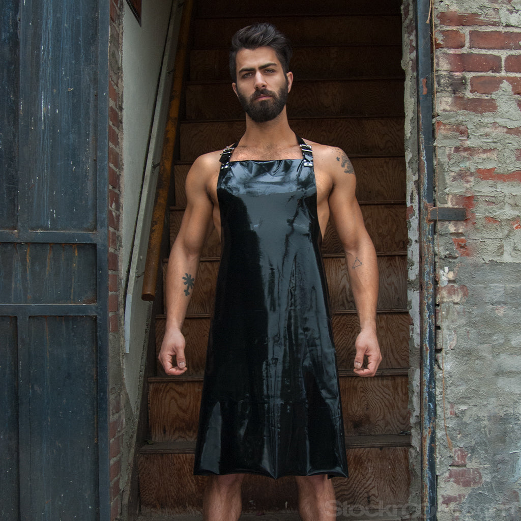 A muscular brunette man with facial hair stands in front of a wooden staircase. He wears the black Rubber Apron by Syren Latex, which has pockets and an adjustable neck strap. The hem of the apron ends at his knees.