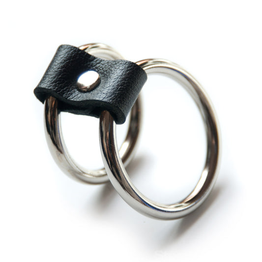 The Double-O Cock Ring, made of two silver metal cock rings lined up next to each other and connected by a small piece of black leather, is displayed against a blank background.
