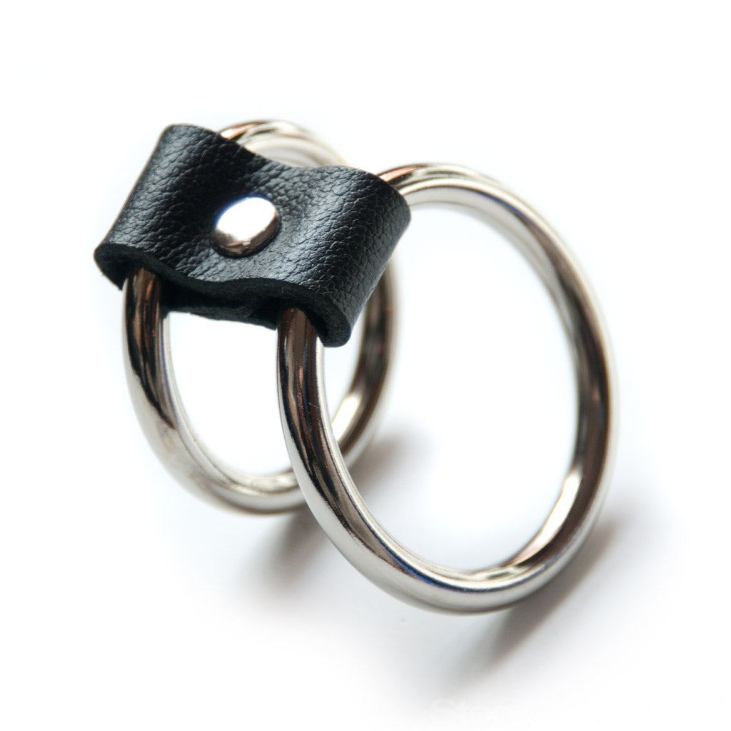 The Double-O Cock Ring, made of two silver metal cock rings lined up next to each other and connected by a small piece of black leather, is displayed against a blank background.