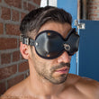 Load image into Gallery viewer, A close-up of a brunette man with light facial hair is shown. He wears the black Mistress Heather Leather Blindfold, which has cupped eye covers with a small metal D-ring between the eyes.