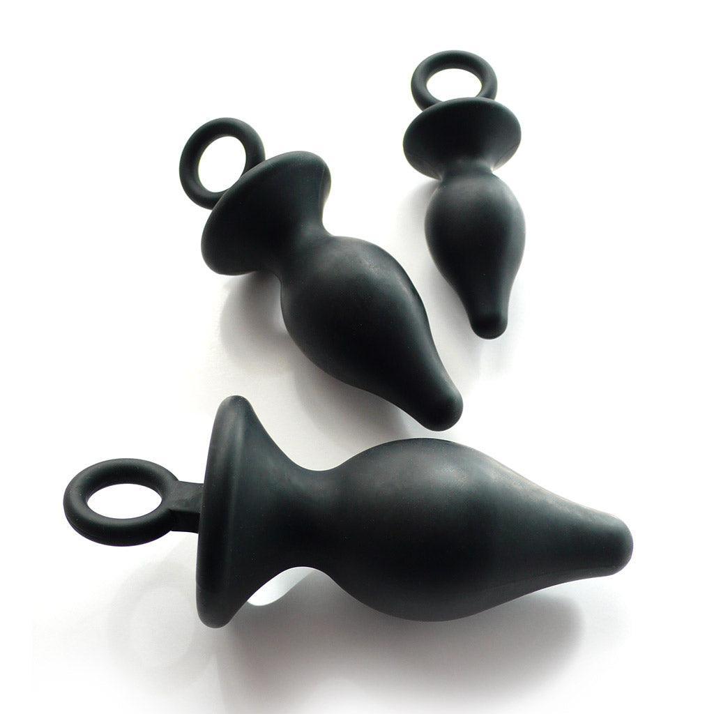 The Anal Trainer Kit is shown against a white background. The kit has a small, medium, and large butt plug, all made of black rubber. The plugs are tapered with thin necks and wide bases at the bottom, and the bases have rings attached to them.