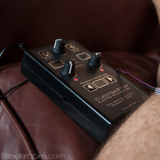 A close-up of an unidentifiable part of a man’s body is shown resting on a brown leather couch. On his leg rests a red silicone pinwheel. The pinwheel is plugged into the Electrastim Sensavox, a grey box with three dials on it and multiple buttons.