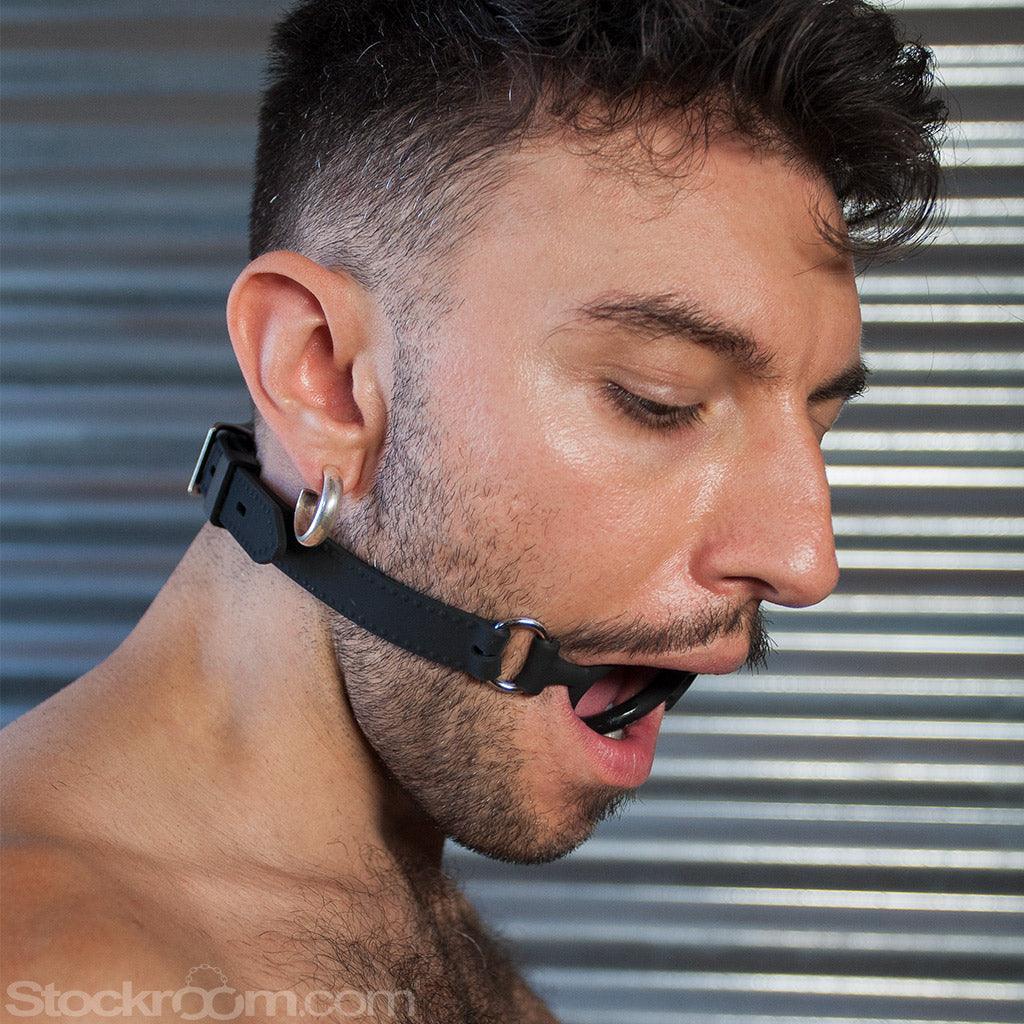 A close-up of a dark-haired man’s face is shown. He has some facial stubble and is wearing a metal hoop earring. He stands in front of a metal wall, looking down, and is wearing the Silicone O-Ring Bondage Mouth Gag.