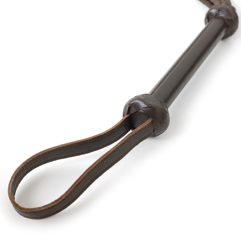 A close-up of the handle of the Brown Leather Braided Whip is displayed against a blank background. The handle widens slightly at the top and the end, where it is surrounded by braided brown leather pieces.