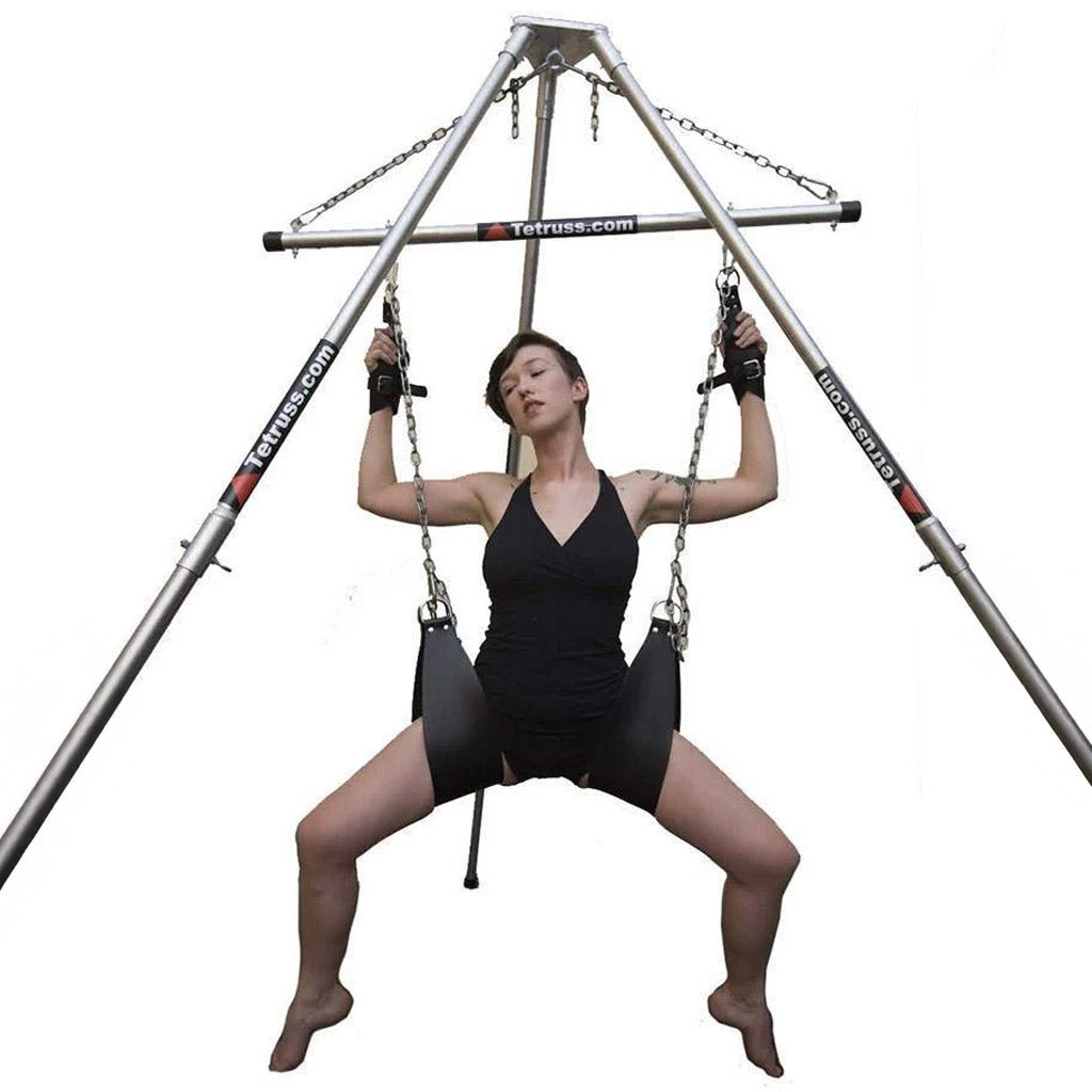 The Tetruss Portable Dungeon Deluxe Bundle is shown in use against a blank background. The spreader bar is connected to the frame, supporting a woman sitting in the leather slings. Her suspension wrist cuffs are attached to the spreader bar.