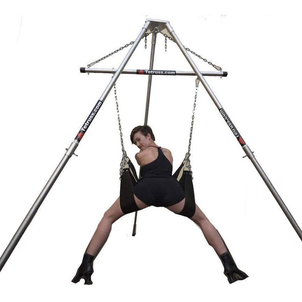 The Tetruss Portable Dungeon Deluxe Bundle is shown in use against a blank background. A woman is shown from the back, leaning forward against one of the stirrups while her legs are supported by the others, all of which are attached to the spreader bar.