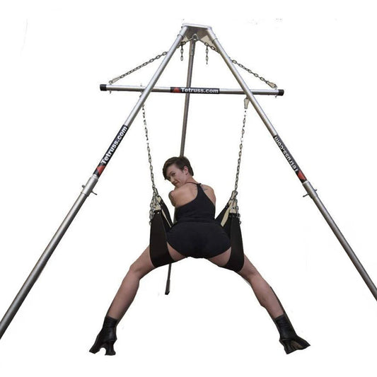 The Tetruss Portable Dungeon Deluxe Bundle is shown in use against a blank background. A woman is shown from the back, leaning forward against one of the stirrups while her legs are supported by the others, all of which are attached to the spreader bar.