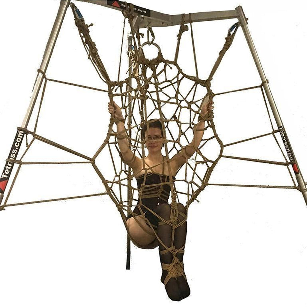 The Tetruss Maxximus Suspension Bondage Frame is shown in use against a blank background. The frame is silver and has 3 poles arranged triangularly, which are connected at the top. A woman is shown tied up in a rope web attached to the frame.