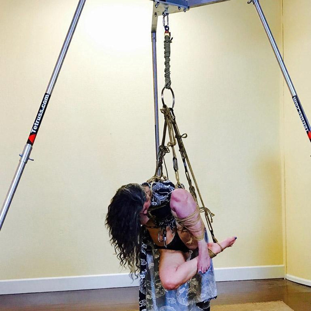 A woman is shown suspended from the Tetruss Maxximus Suspension Bondage Frame. She is suspended by her leg, which is tied in a bent position. Her brown hair hands down as she lowers her head.