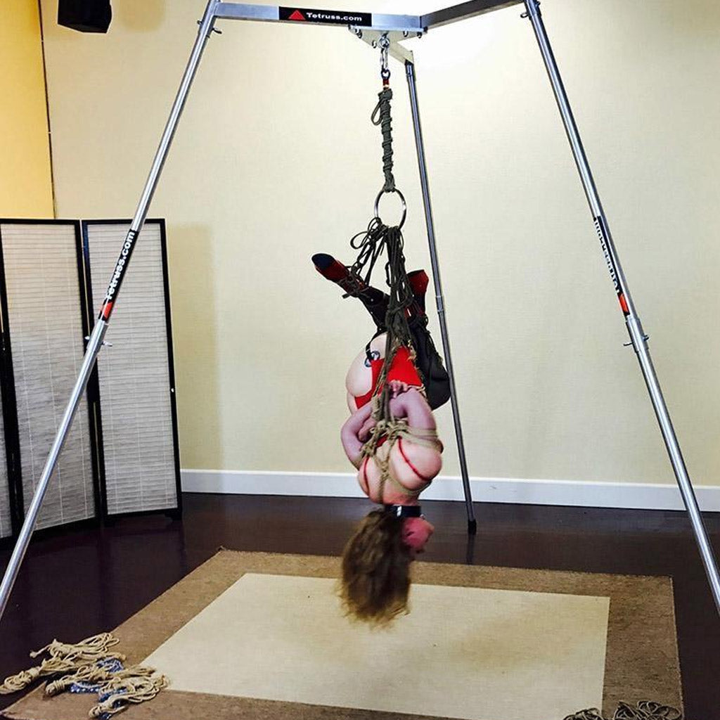 A woman is suspended from the Tetruss Maxximus Suspension Bondage Frame. Her arms are tied behind her back, and she is suspended upside down by them. She is wearing a red bodysuit and long black boots. Her face is visibly red.