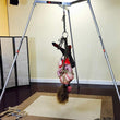 Load image into Gallery viewer, A woman is suspended from the Tetruss Maxximus Suspension Bondage Frame. Her arms are tied behind her back, and she is suspended upside down by them. She is wearing a red bodysuit and long black boots. Her face is visibly red.