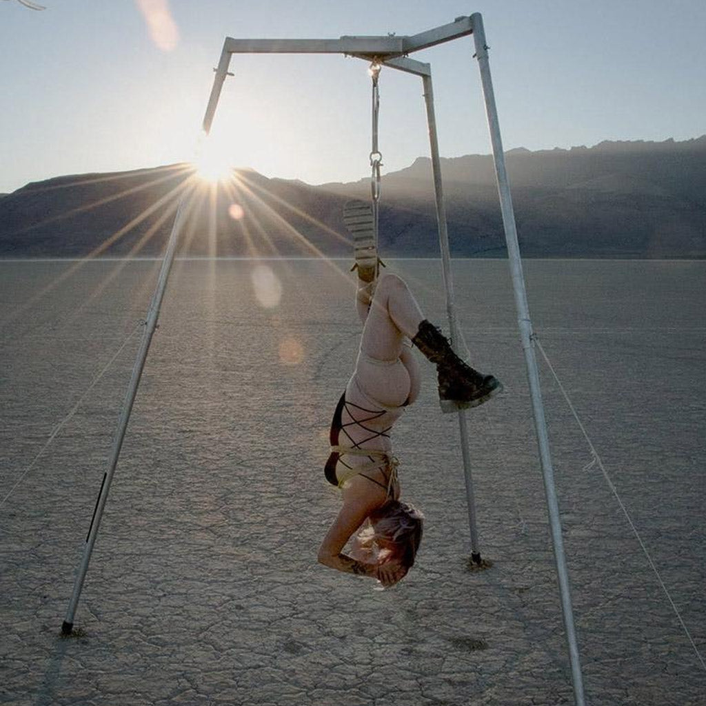 A woman is shown suspended from the Tetruss Maxximus Suspension Bondage Frame. She is outdoors in the desert and is hanging upside down, suspended by ropes tied around her torso.