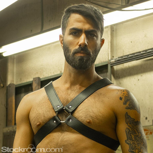 A brunette man with facial hair is shown from the waist up, standing in an industrial room. He wears the black Leather Chest. A strap goes over each shoulder and under his arms. They are attached in the middle to a metal O-ring.