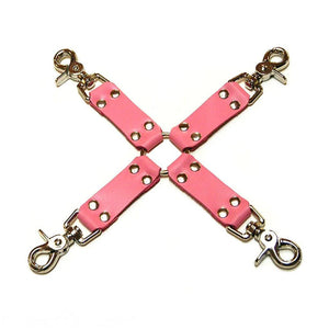The pink Leather Hog Tie is displayed against a blank background. A metal O-ring is in the center with four leather strips attached to it in the shape of an X. Each leather strip has a metal snap hook on the end.