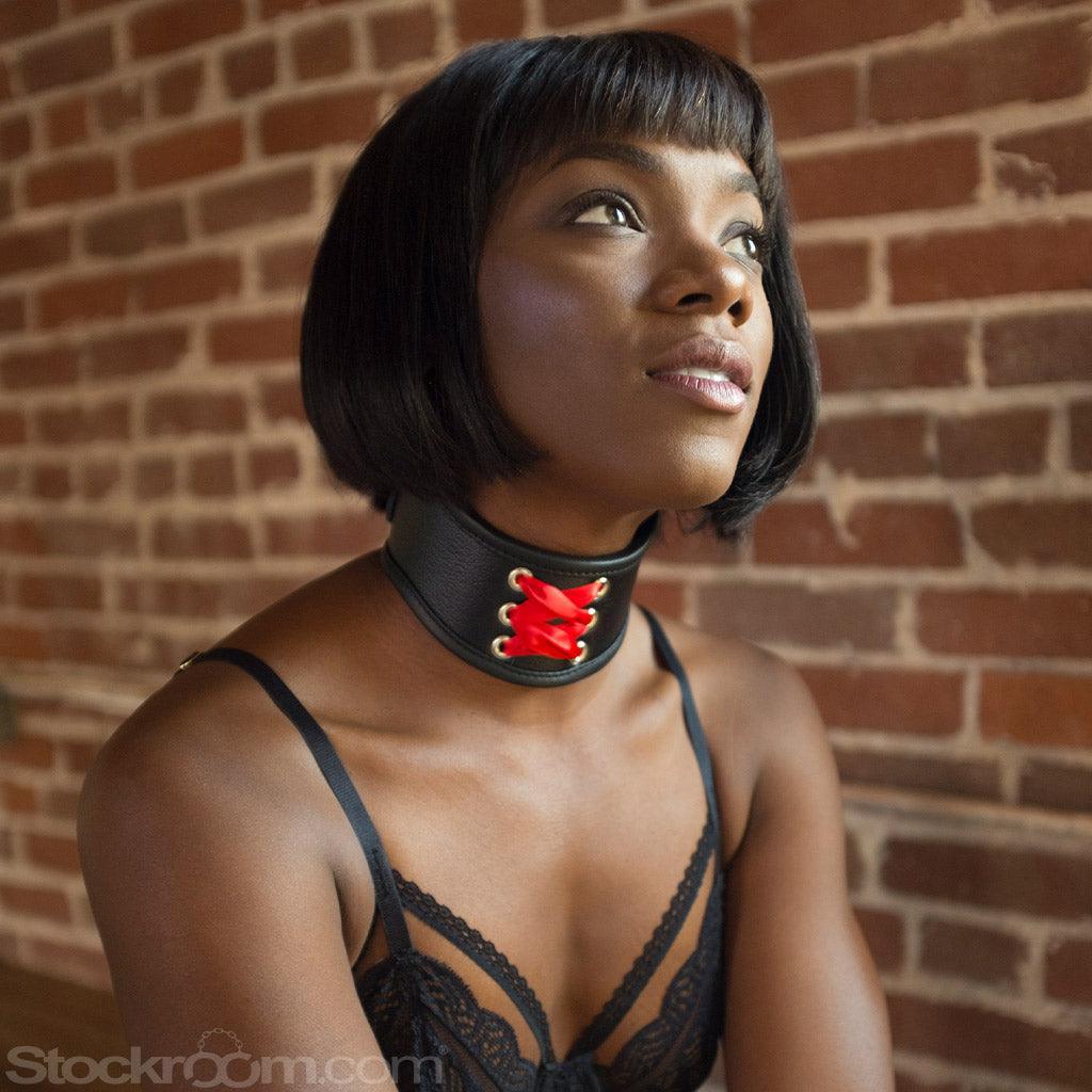 A woman with a black bob wearing black lingerie kneels in front of a brick wall. She wears the Red Laced BDSM Posture Collar, a small posture collar made of black leather with decorative red corset lacing on the front.