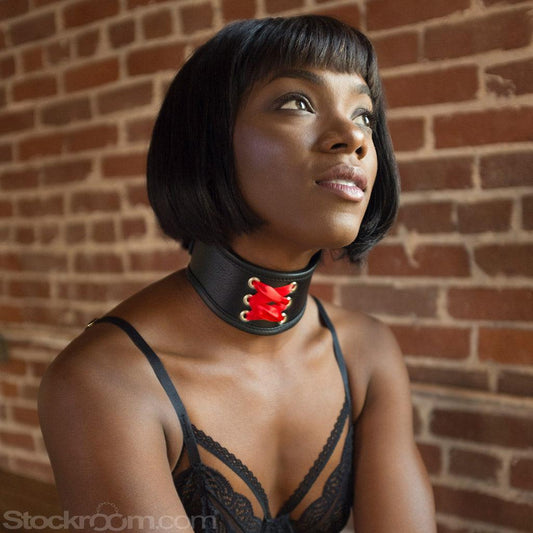 A woman with a black bob wearing black lingerie kneels in front of a brick wall. She wears the Red Laced BDSM Posture Collar, a small posture collar made of black leather with decorative red corset lacing on the front.