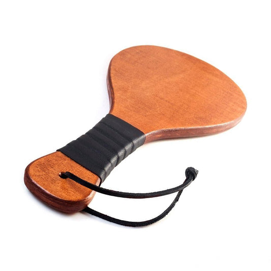 The Leather Wrapped Round Wood Spanking Paddle is shown against a blank background. The paddle is made of light-colored wood and has a black wrist loop at the bottom. The middle of the handle is wrapped in a black leather strip.