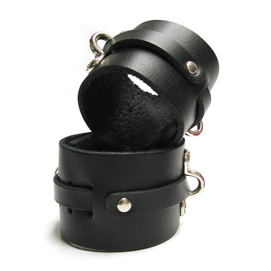 The KinkLab Bondage Basics Leather Wrist Cuffs are shown against a blank background. They are made of a wide piece of black leather with a thin piece of leather that wraps around them, through notches and under metal D-rings.