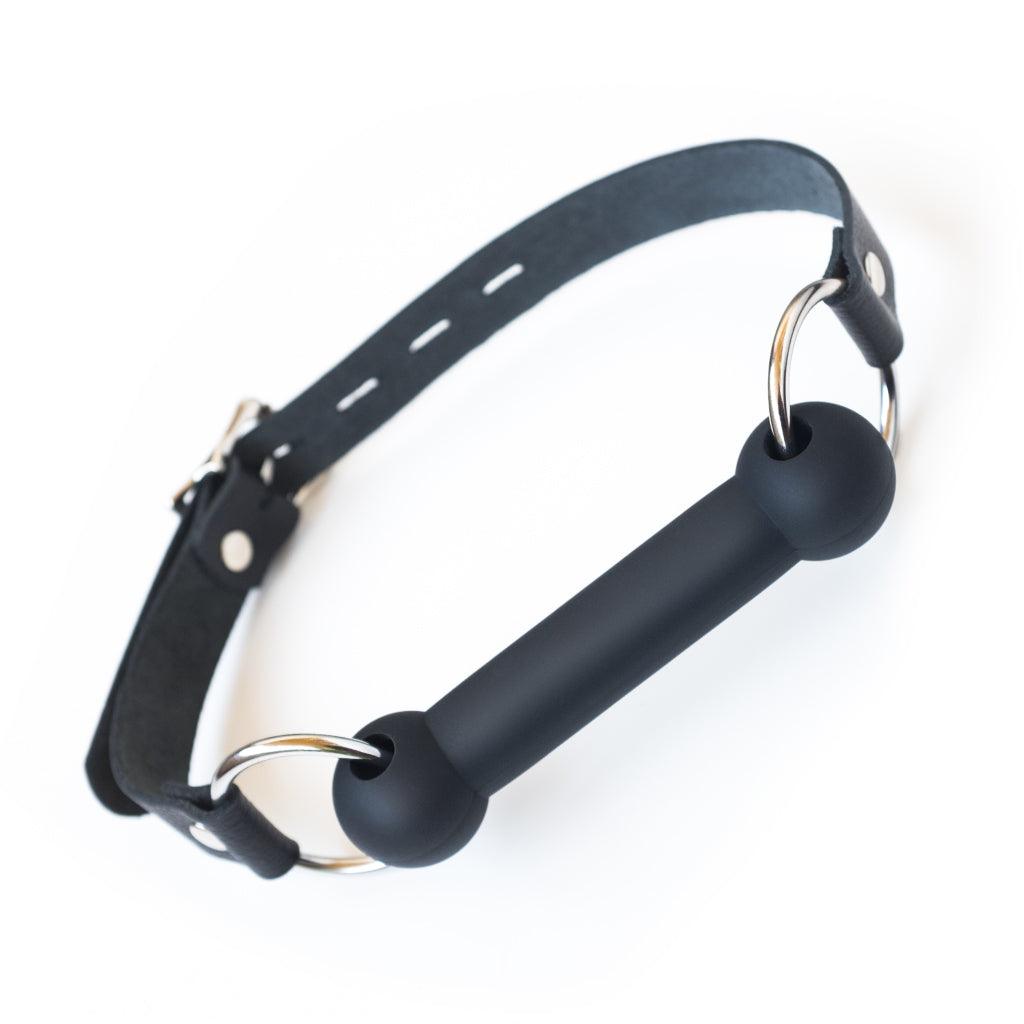 The Kinklab Silicone Bit Gag With A Leather Strap is shown against a blank background. The gag bit is made of matte black silicone and attached to the leather head strap with metal O-rings.