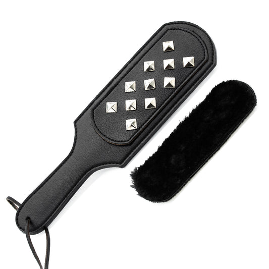 A square leather paddle with square, spiky studs rests on a white background. Next to it is a black, fuzzy attachment.