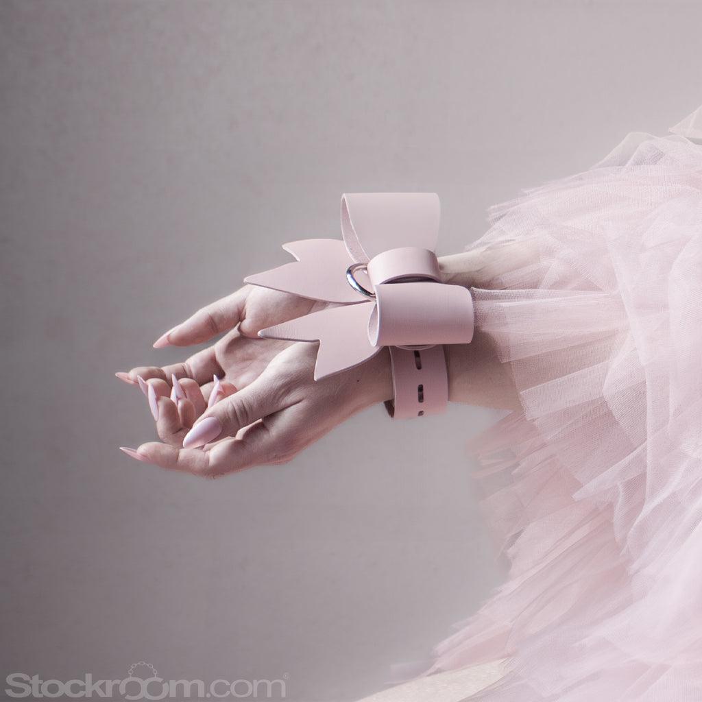 Two feminine hands with long, pink nails extend from the right edge of the picture. They're held together with light pink leather straps, tied in a bow..