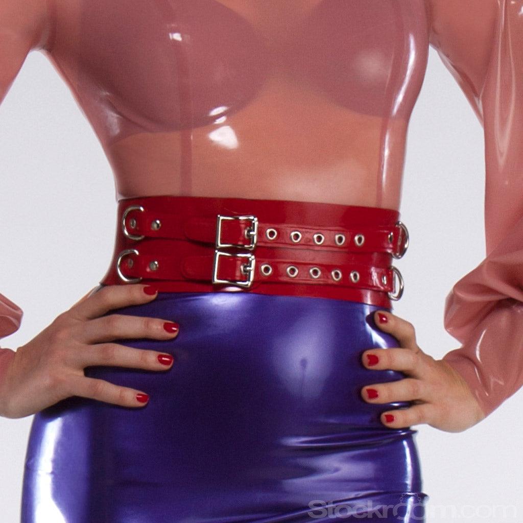 A close-up of a woman's waist is shown in front of a grey background. Over her pink latex blouse and purple latex skirt, she wears the latex waist cuff in red by Syren Latex. The cuff has two buckles in the front and has D-rings on the sides.
