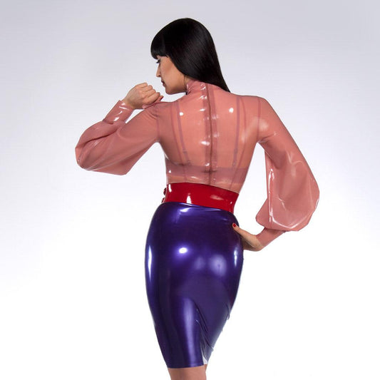 A woman with dark hair and red lipstick wearing a pink latex blouse and purple latex skirt poses in front of a grey backdrop, facing away from the camera. She wears a red latex Waist Cuff by Syren Latex.